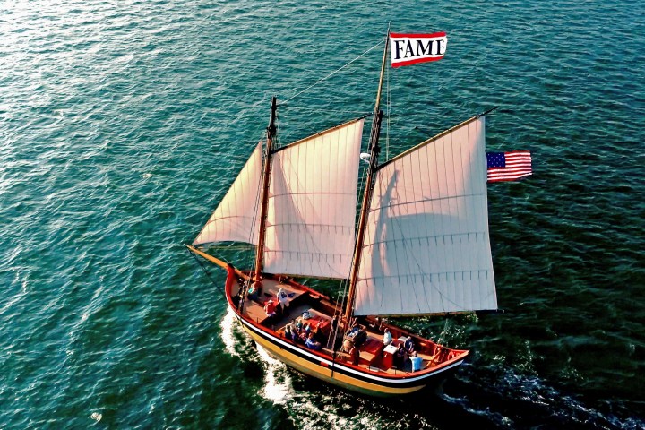 Aerial view of sailboat with 'FAME' flag sailing on water, American flag visible, in sunny weather.