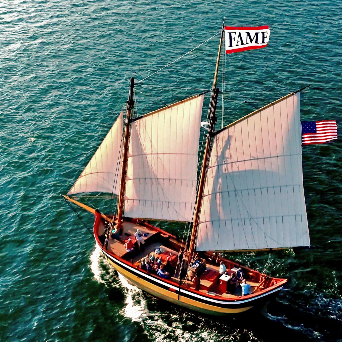 Aerial view of sailboat with 'FAME' flag sailing on water, American flag visible, in sunny weather.