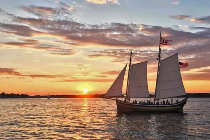 Sailing ship with flags at sunset on open water with an orange sky and distant horizon.