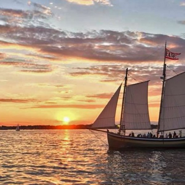 Sailing ship with flags at sunset on open water with an orange sky and distant horizon.