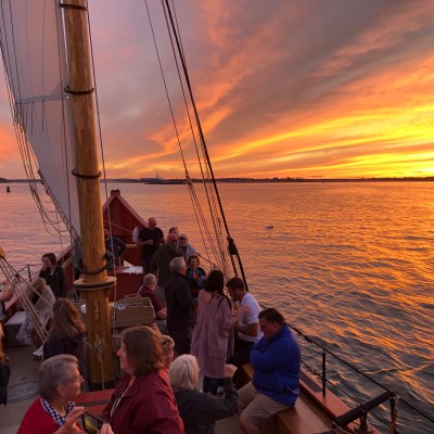 People on a sailboat at sunset with a vibrant orange sky over the water.