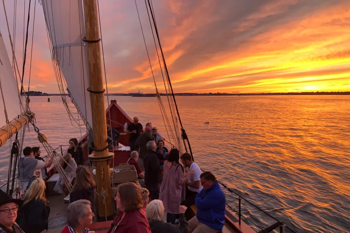 People on a sailboat at sunset with a vibrant orange sky over the water.