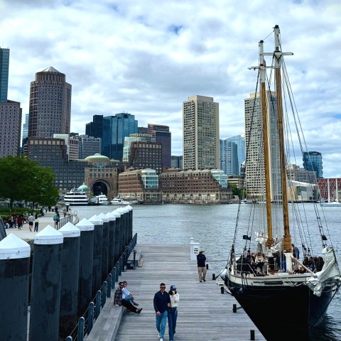 Docked sailboat with city skyline and trees in the background under a cloudy sky.