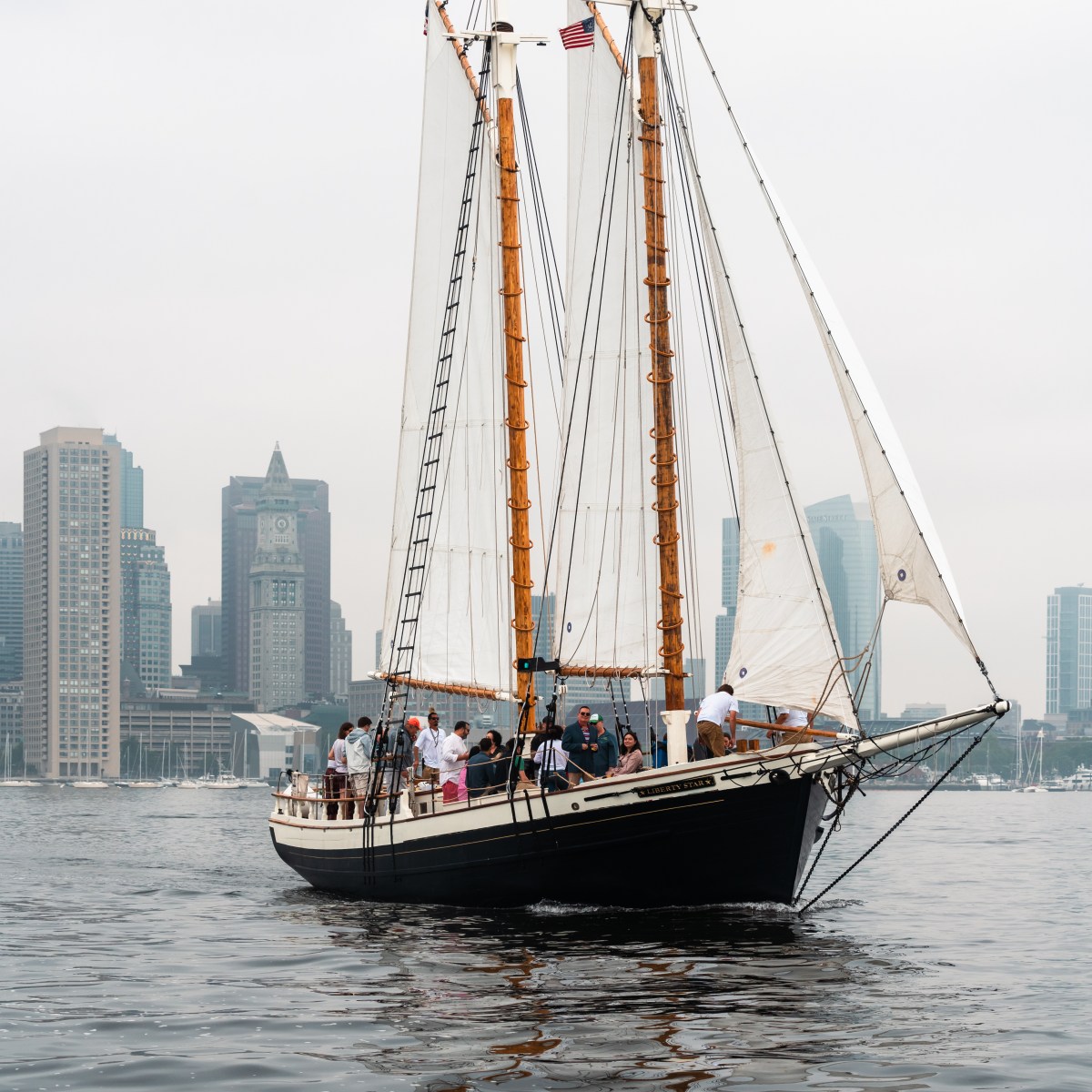 Sailboat with people in city harbor, tall buildings in background, cloudy sky.