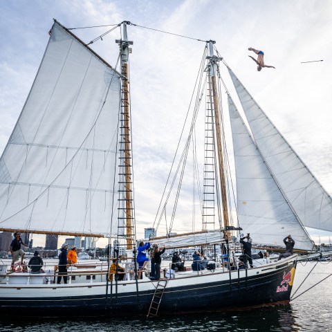 Person diving from a tall ship with white sails, city skyline in the background.