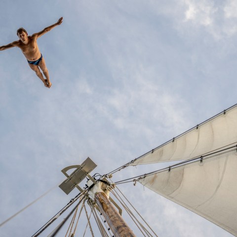 Person diving from a sailboat mast, blue sky background, sails visible on right.