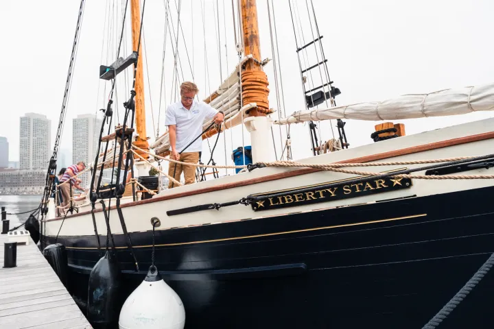 Two people on a docked sailboat named Liberty Star, with city skyline in the background.