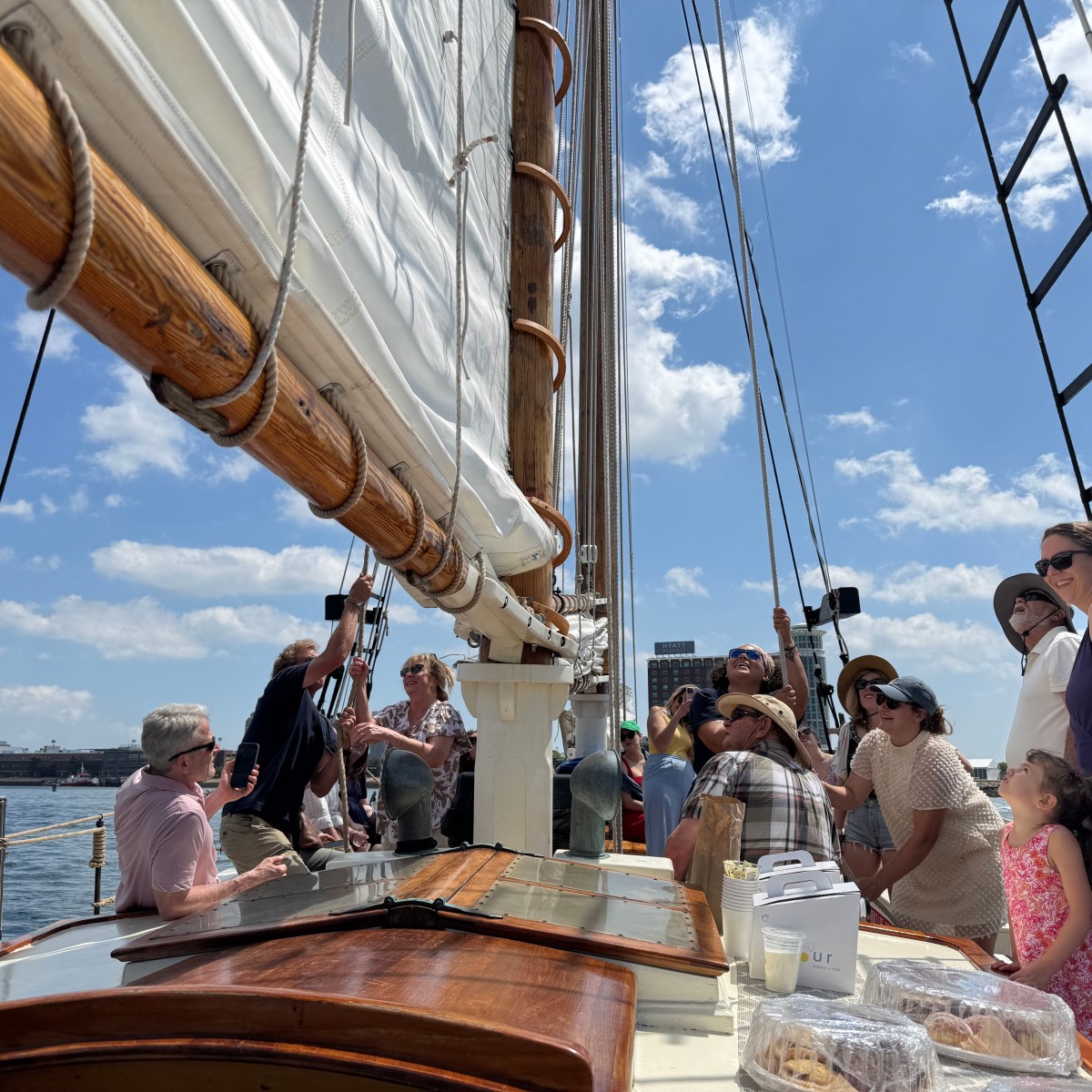 Group of people on a sailboat deck under a clear sky, with visible sails and mast.
