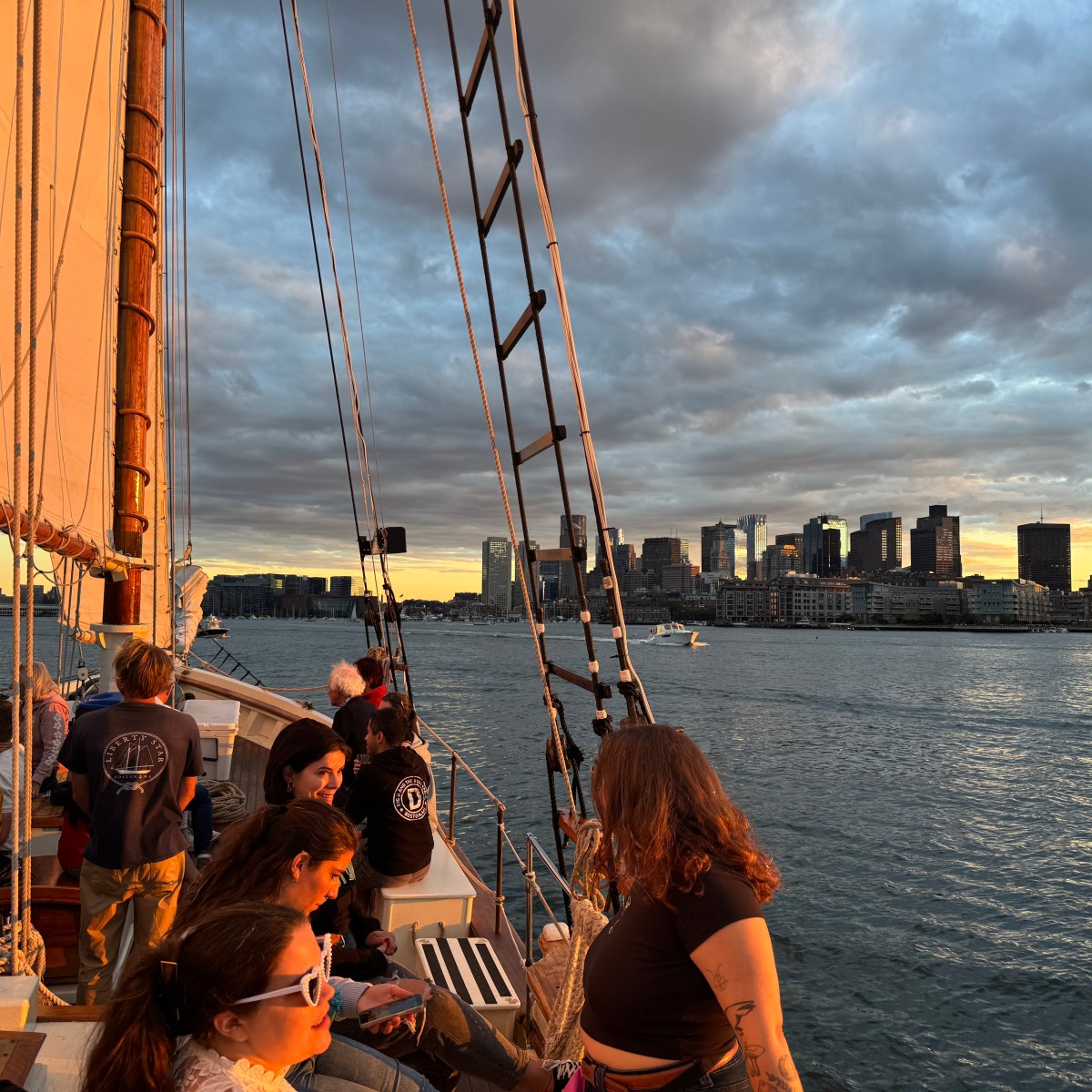 People on a sailboat at sunset with a city skyline in the background.