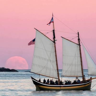 Sailboat with full sails on calm sea at sunset, large moonrise in background.