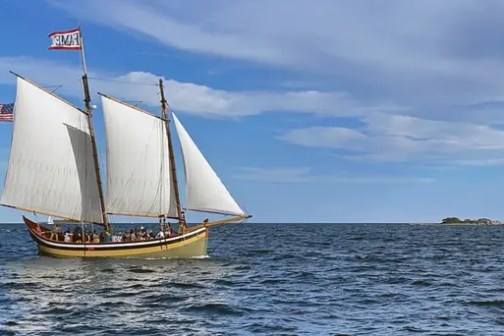 A sailboat with two sails and flags on a large body of water under a blue sky with clouds.